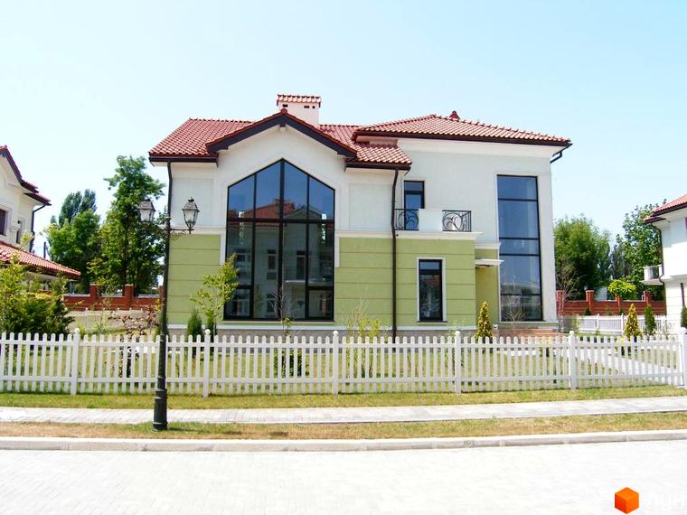 Зображення двоповерхового будинку в Котеджному містечку Аріадна з великими вікнами та червоною черепицею. Перед будинком білий паркан та доглянутий газон.