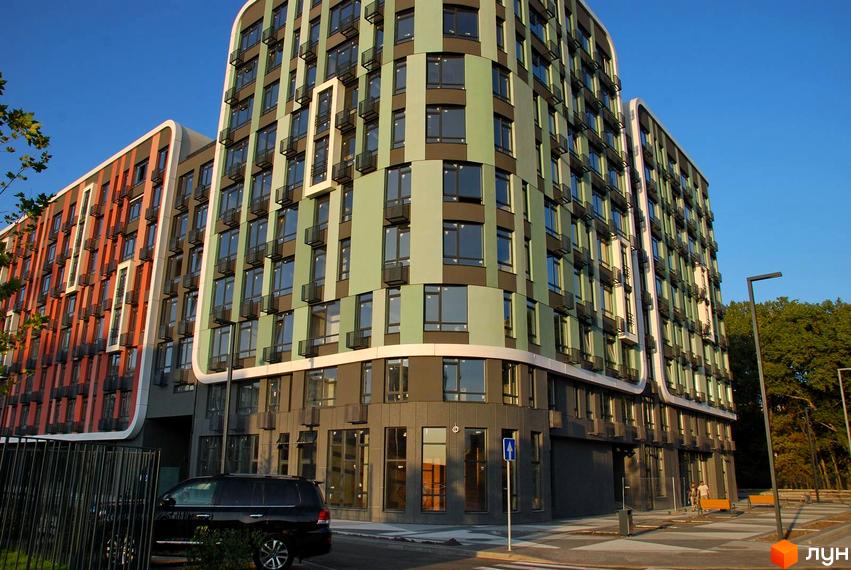 Фасад житлового комплексу Respublika з зеленими та помаранчевими панелями, великими вікнами та балконами. Поруч дорога, припаркований автомобіль та дерева.