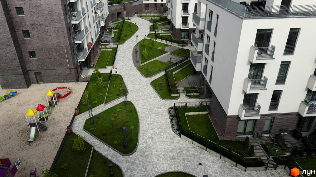 Зовнішній вигляд новобудови ЖК Villa Magnolia з білими фасадами та балконами. Прибудинкова територія з пішохідними доріжками, зеленими зонами та дитячим майданчиком.