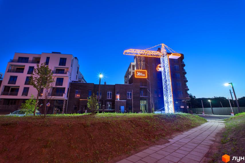 Житловий комплекс Villa Magnolia з підсвіченим краном та фасадами будівель у вечірній час. Територія навколо комплексу з доріжками та зеленими насадженнями.