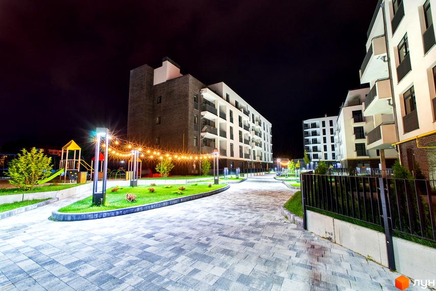 Житловий комплекс Villa Magnolia з фасадами білого кольору та балконами. Територія освітлена гірляндами та ліхтарями, є дитячий майданчик.