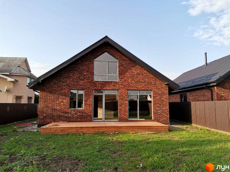 Котеджне містечко КМ Баварське село: цегляний будинок з великими вікнами та дерев'яною терасою на тлі блакитного неба. Навколо будинку зелена галявина та паркан.