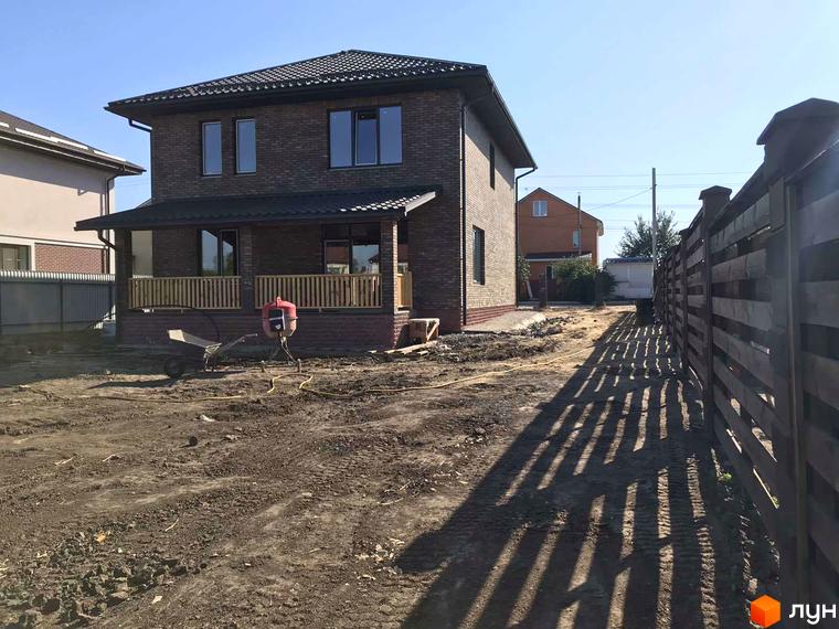 Котеджне містечко English village: двоповерховий цегляний будинок з дерев'яною верандою та огорожею на тлі блакитного неба. Територія навколо будинку вкрита землею, видно будівельні матеріали та паркан.