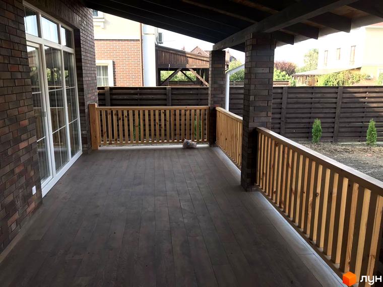 Тераса з дерев'яними перилами та підлогою з темних дощок у котеджному містечку КМ English village. На задньому плані паркан та зелені насадження.