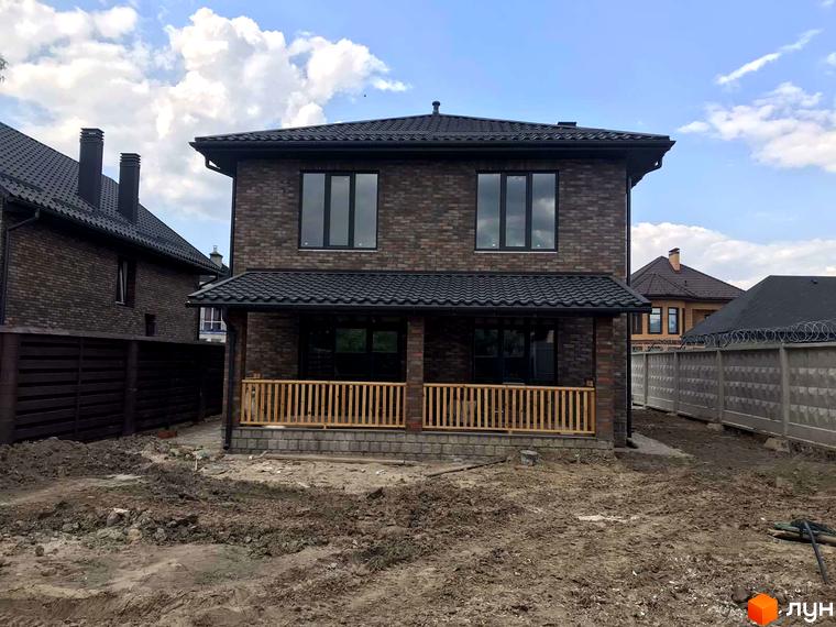 Котеджне містечко English village: двоповерховий будинок з цегляним фасадом та чорним дахом, перед будинком веранда з дерев'яною огорожею та ділянка з будівельним майданчиком.