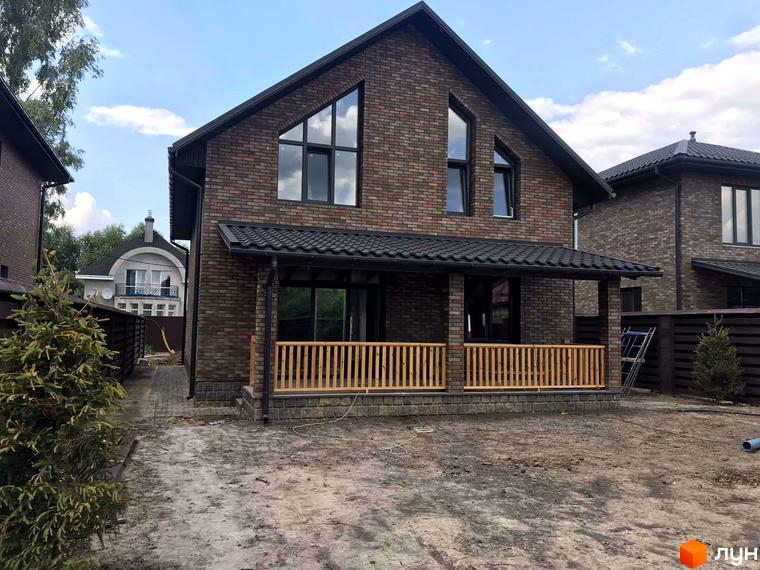 Котеджне містечко English village: двоповерховий цегляний будинок з коричневим дахом, верандою та дерев'яною огорожею. Навколо будинку облаштована прибудинкова територія.