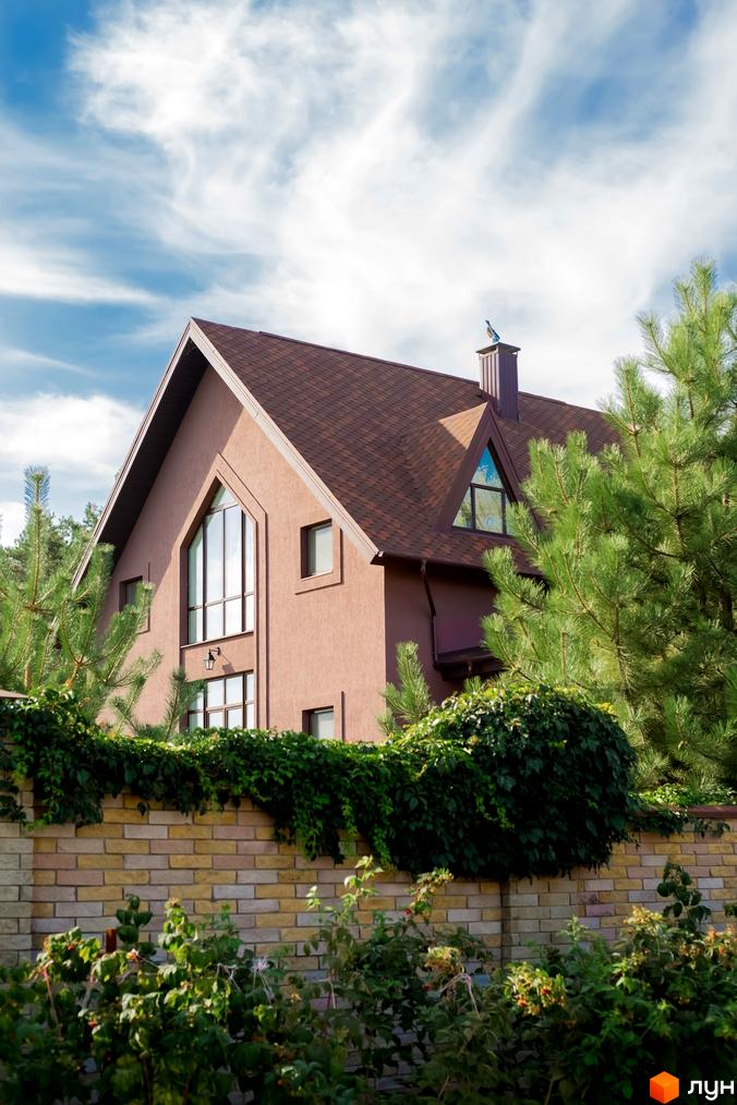 Котеджне містечко КМ Барвіта. Зовнішній вигляд будинку з коричневою черепицею та великими вікнами, оточений зеленню та цегляним парканом.