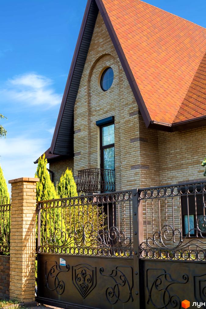 Котеджне містечко КМ Barvita: кам'яний фасад будинку з червоним дахом та кованою огорожею. Зелені туї та блакитне небо.