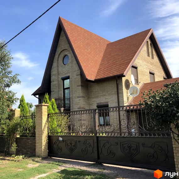 Котеджне містечко КМ Барвіта: цегляний будинок з червоним дахом та кованими воротами. Навколо будинку зелені насадження та паркан.