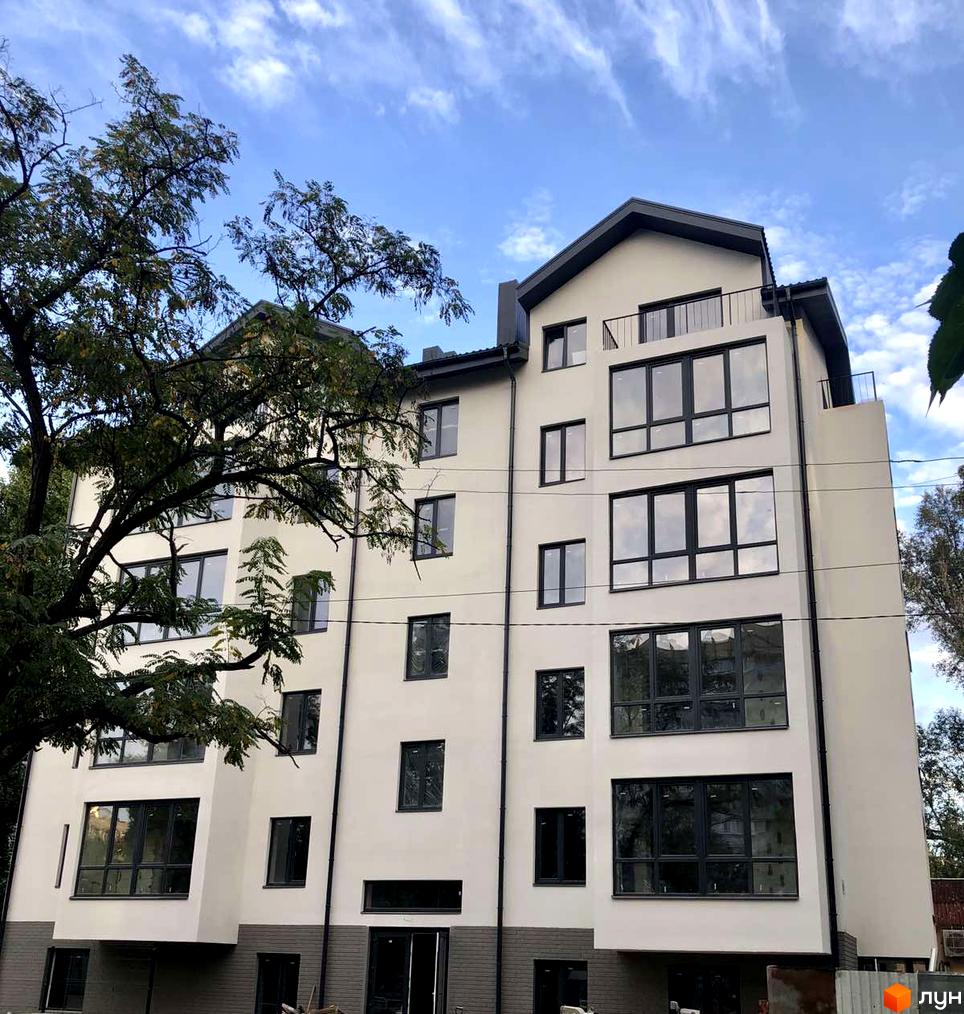 Зовнішній вигляд клубного будинку «Зразковий» з великими вікнами та балконами на тлі блакитного неба з хмарами. Навколо будівлі видно зелені дерева.