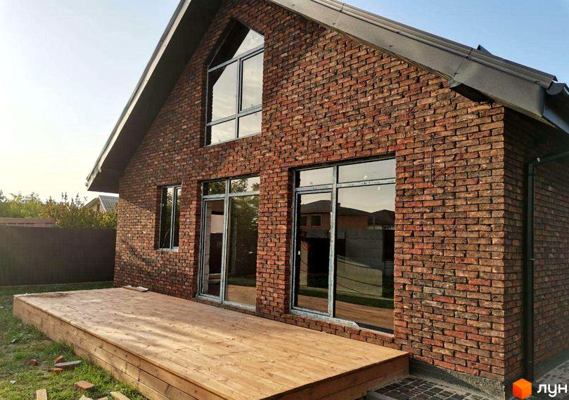 Котеджне містечко Баварське село: цегляний будинок з великими вікнами та дерев'яною терасою. Навколо будинку зелена трава та паркан.