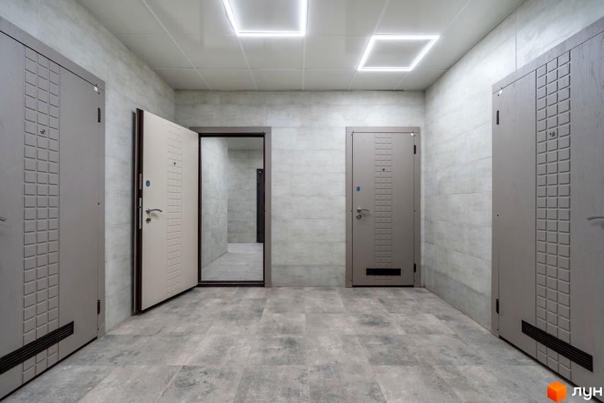 Вхідна група житлового комплексу на вул. Ломаківська (Мічуріна), 19б. Сірі двері з декоративними елементами та світлі двері, що відчинені, ведуть у коридор з плиткою на стінах та підлозі.