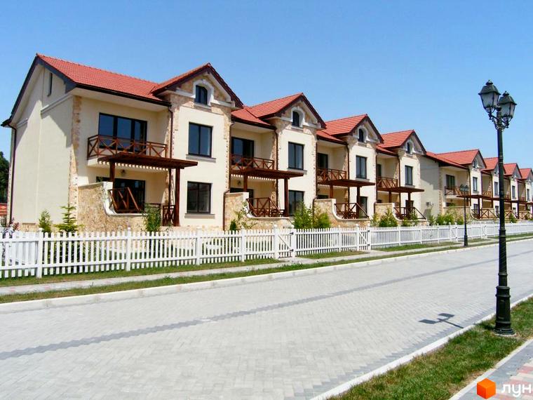 Фото Котеджного містечка Аріадна: двоповерхові будинки з балконами та червоними дахами, огороджені білим парканом. Перед будинками – тротуарна плитка та газон.