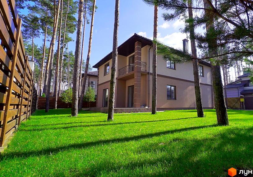 Котеджне містечко КМ Сонячний 2. Двоповерховий будинок з балконом та коричневим дахом, розташований серед високих сосен на доглянутій зеленій ділянці.