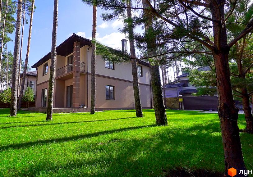 Котеджне містечко КМ Сонячний 2. Двоповерховий будинок з балконом та коричневим дахом, оточений високими соснами та доглянутим зеленим газоном.