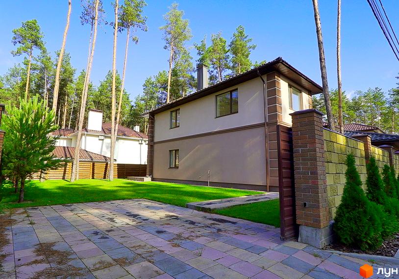 Котеджне містечко КМ Сонячний 2. Зовнішній вигляд двоповерхового будинку з коричневим дахом та світло-бежевими стінами, огородженого цегляним парканом. Прибудинкова територія з бруківкою та зеленим газоном.