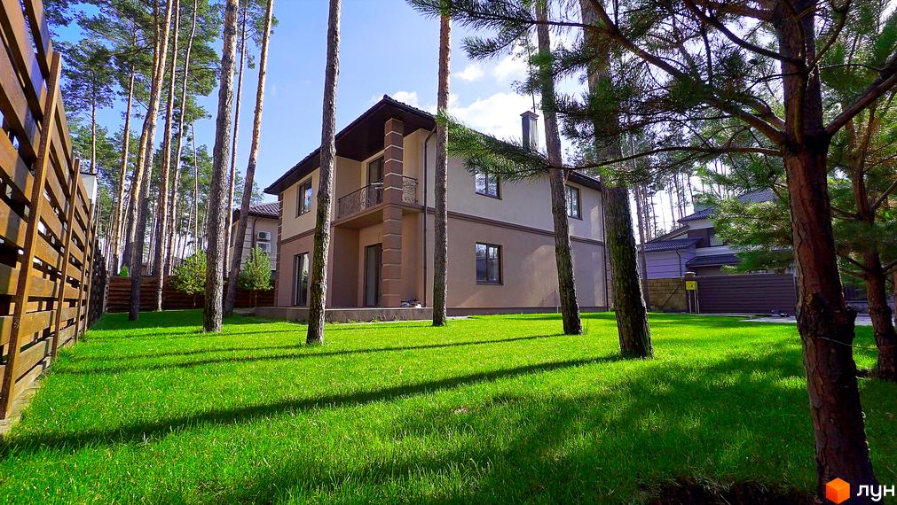 Котеджне містечко КМ Сонячний 2. Двоповерховий будинок з балконом та коричневим дахом на тлі зеленого газону та сосен.