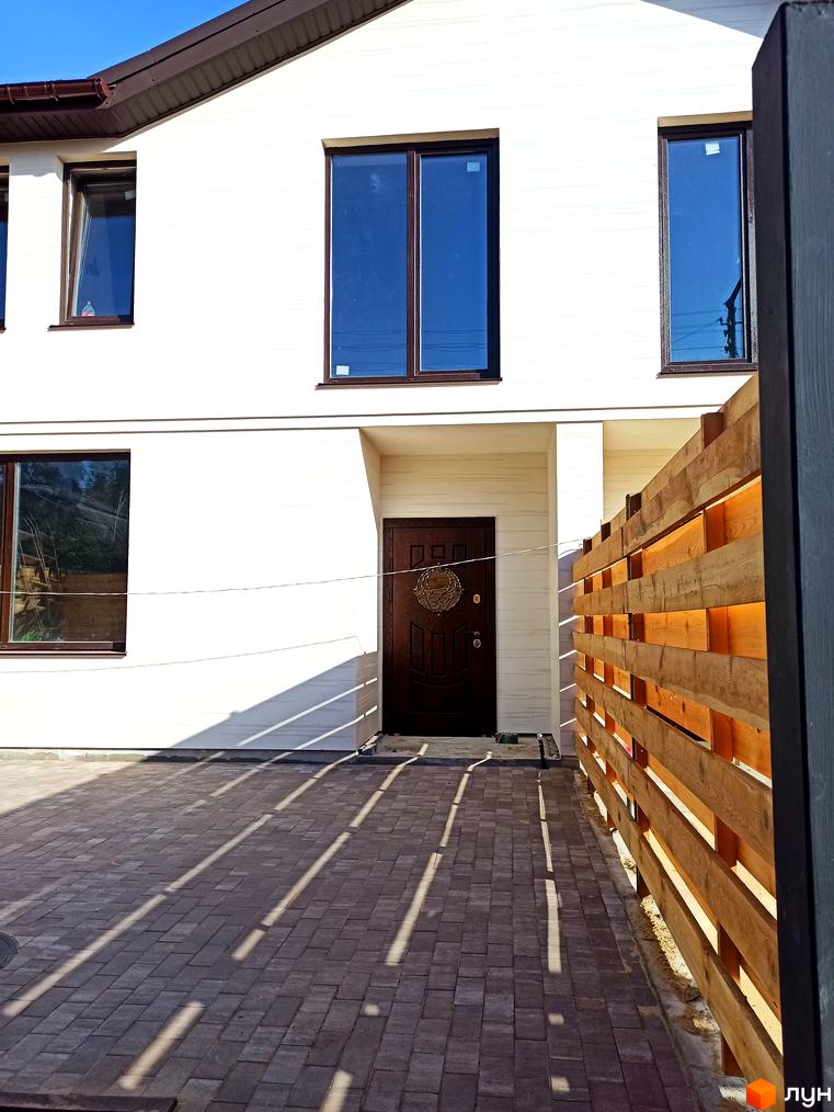 Білий фасад таунхауса Travertino з коричневими віконними рамами та дверима. На подвір'ї укладена бруківка, поруч дерев'яний паркан.