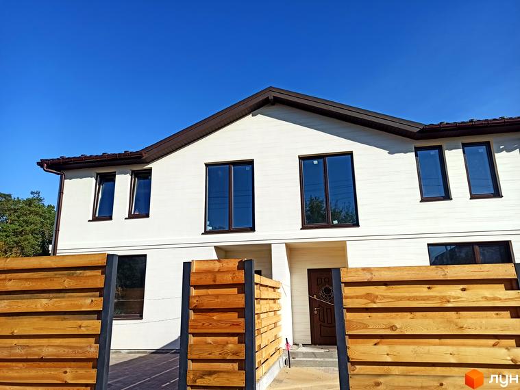 Двоповерховий таунхаус Travertino зі світлим фасадом, коричневим дахом та темними вікнами, перед яким розташований дерев'яний паркан.