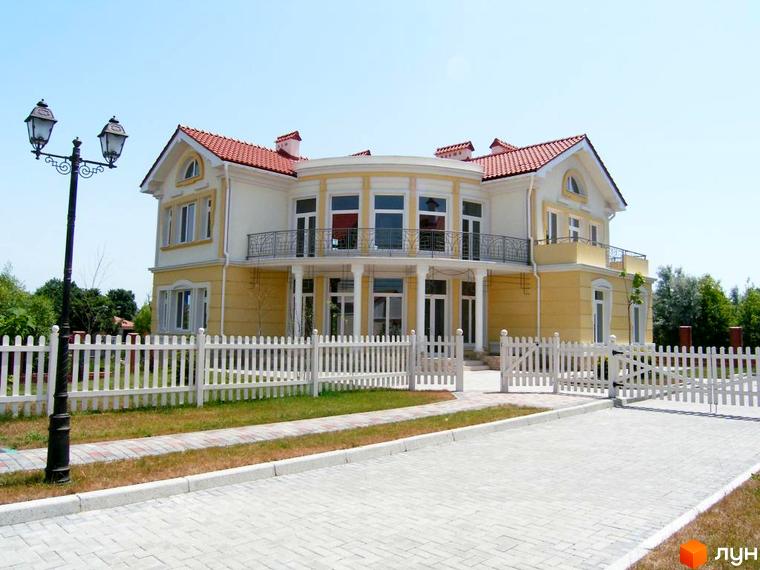 Зображення двоповерхового будинку в Котеджному містечку Аріадна з червоним дахом та білим парканом. Перед будинком бруківка та газон.