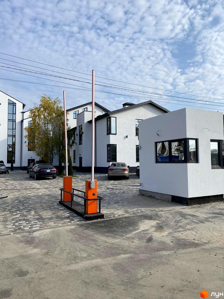 Зовнішній вигляд будинків житлового комплексу Aura з парканом та шлагбаумом. На території комплексу розташовані автомобілі та дерева.