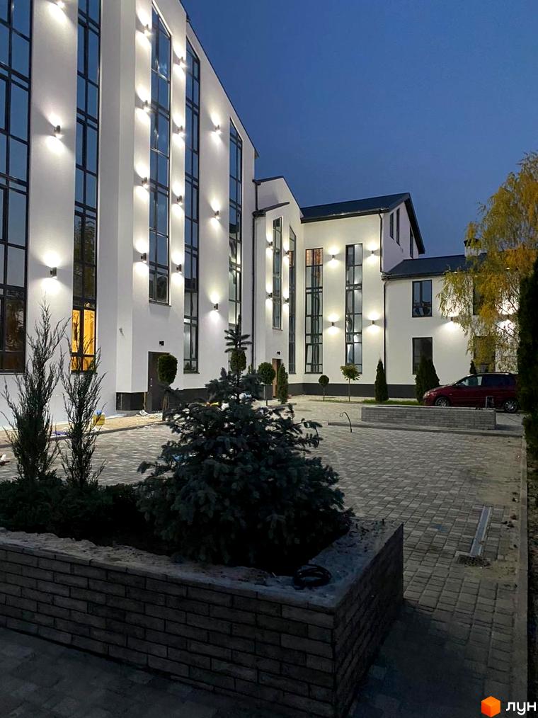 Зовнішній вигляд новобудови ЖК Aura з підсвіченими фасадами та вікнами, прибудинкова територія з бруківкою та деревами.
