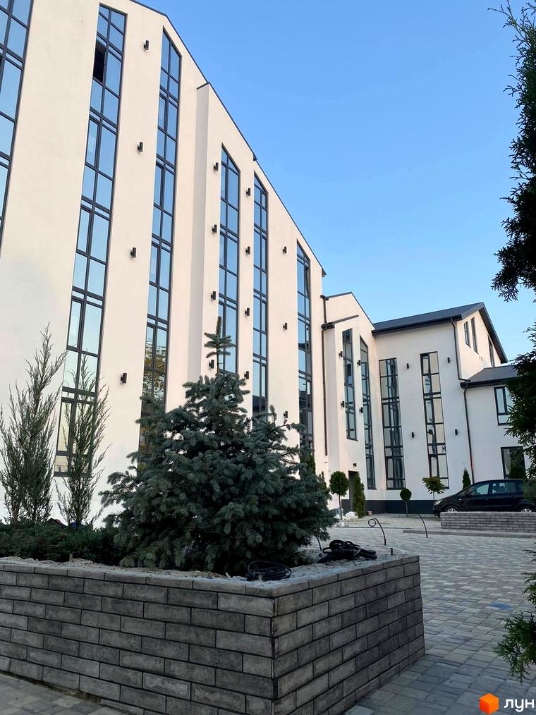 Зовнішній вигляд житлового комплексу Aura з білими фасадами та великими вікнами. Прибудинкова територія з зеленими насадженнями та бруківкою.