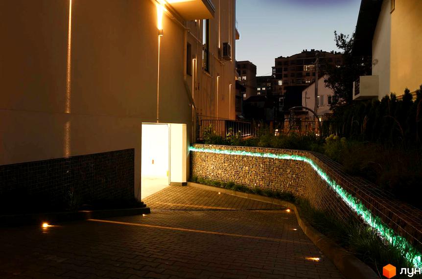 Житловий комплекс Baltiysʹka Hall ввечері з освітленим фасадом та підсвіченою зеленою смугою на габіонній стіні. Бруківка з наземними ліхтарями.