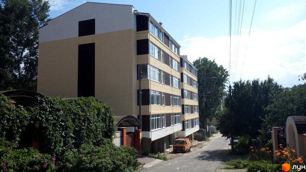 Житловий комплекс Будинок на Бестужева – це нова багатоповерхова будівля з світло-бежевим фасадом та темними елементами. Навколо комплексу зелена зона з деревами та кущами.