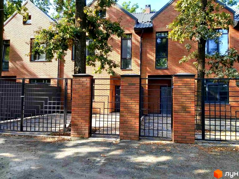 Таунхаус Brighton Lisova з червоної цегли, з металевою огорожею та кованими воротами. Навколо будинку зелені дерева та доглянута прибудинкова територія.
