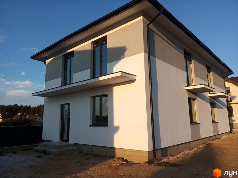 Котеджне містечко КМ Сосновий маєток: двоповерховий будинок з сірим та білим фасадом, вікнами та балконами. Навколо будинку видно ділянку з піском та зеленню.