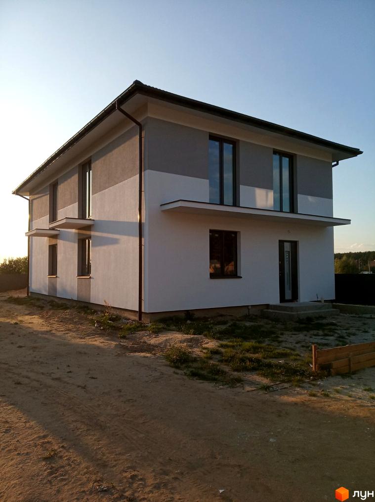 Двоповерховий будинок Котеджного містечка Сосновий маєток з біло-сірим фасадом та темними вікнами. Навколо будинку ґрунтова ділянка з невеликою кількістю трави.