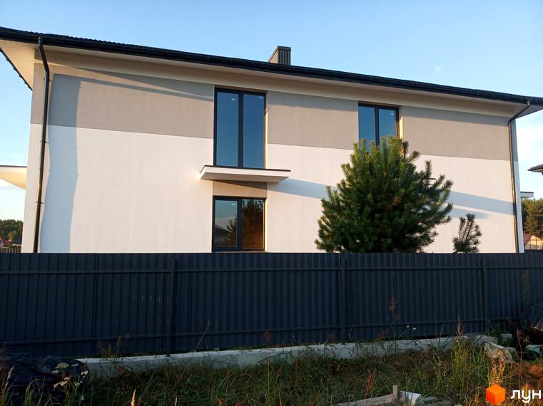 Фасад двоповерхового будинку КМ Сосновий маєток з білими та сірими стінами, темними вікнами. Перед будинком видно зелене дерево та паркан.