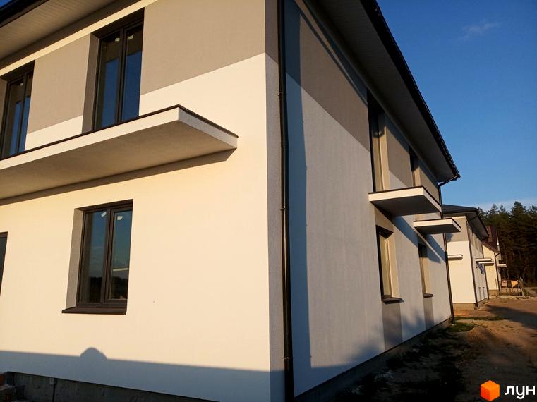 Будинок Котеджного містечка Сосновий маєток з світлими та сірими фасадами, темними вікнами та виступами. Поруч ґрунтова дорога та ліс.