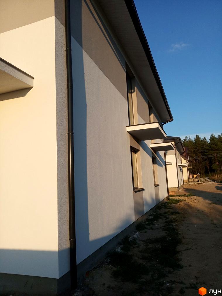 Фасад будинку Котеджного містечка Сосновий маєток: білі та сірі стіни, темні водостічні труби. Поруч ґрунтова дорога, сосновий ліс.