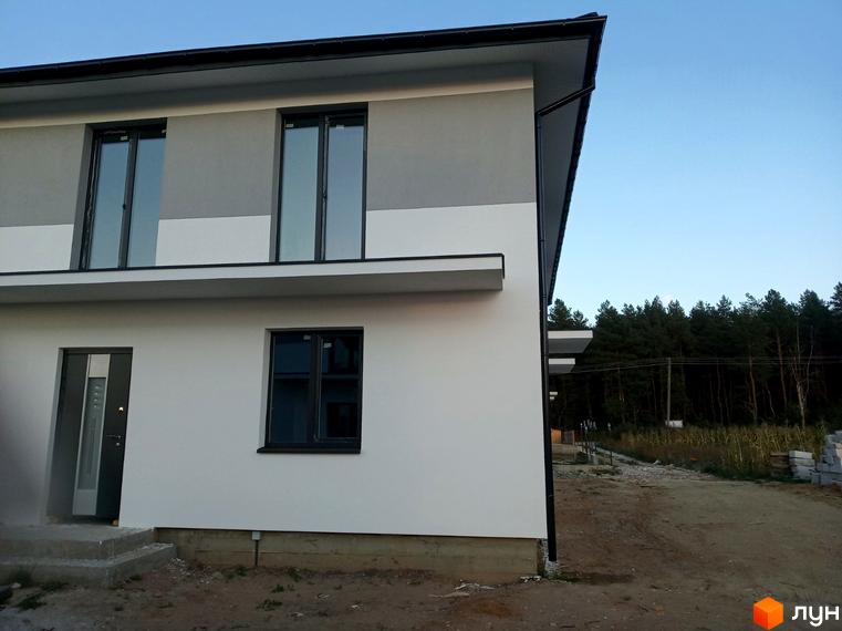 Двоповерховий будинок Котеджного містечка Сосновий маєток з біло-сірим фасадом та темними вікнами. Навколо ґрунтова ділянка та ліс.