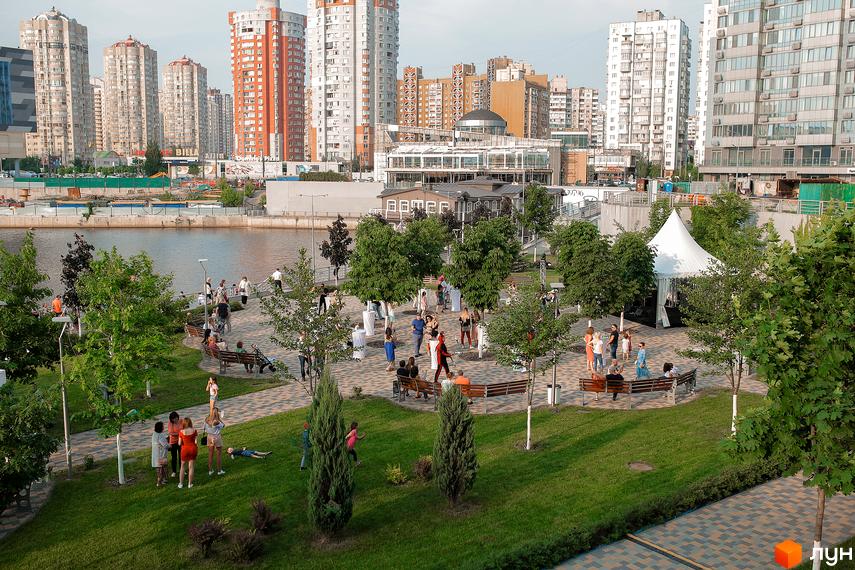 Житловий комплекс RiverStone з високими будинками та зеленою прибудинковою територією біля річки. Люди відпочивають на газоні та біля води.