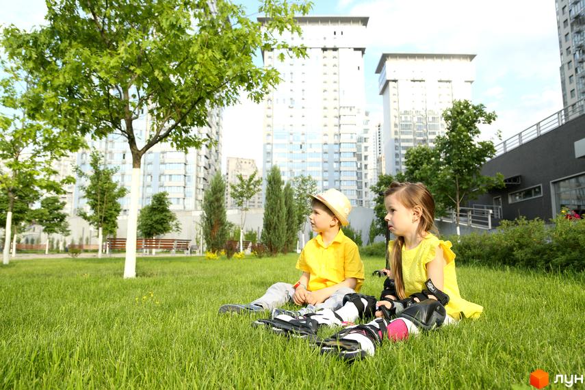 Діти в захисному спорядженні сидять на траві біля багатоповерхових будинків ЖК RiverStone. Навколо зелені дерева та кущі, що створює приємну атмосферу для відпочинку.