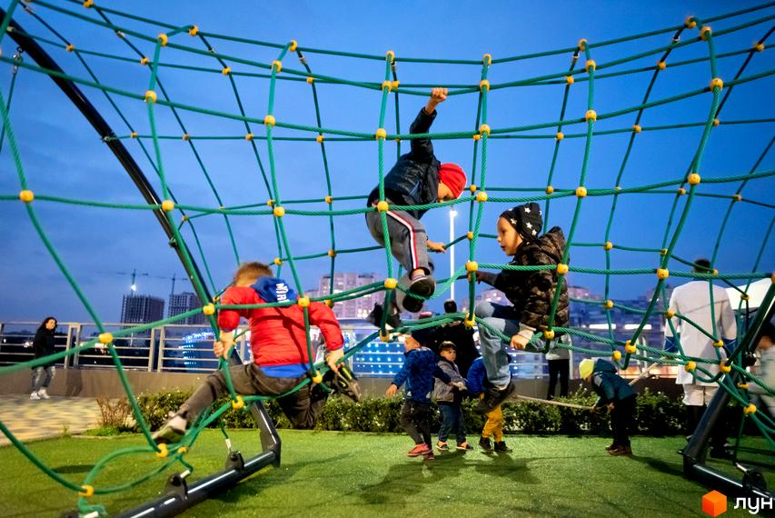 Діти граються на мотузковому атракціоні на території житлового комплексу RiverStone. На задньому плані видно міські будівлі та вечірнє небо.