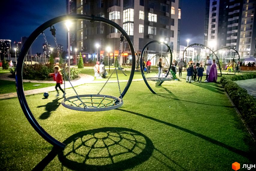 Житловий комплекс RiverStone: дитячий майданчик з гойдалками та дітьми ввечері. Навколо зелена трава, дерева та освітлені багатоповерхові будинки.