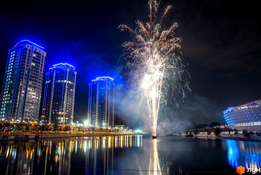 Житловий комплекс RiverStone з синьою підсвіткою на фоні нічного неба та феєрверку. Будинки розташовані біля річки, яка відбиває вогні міста.