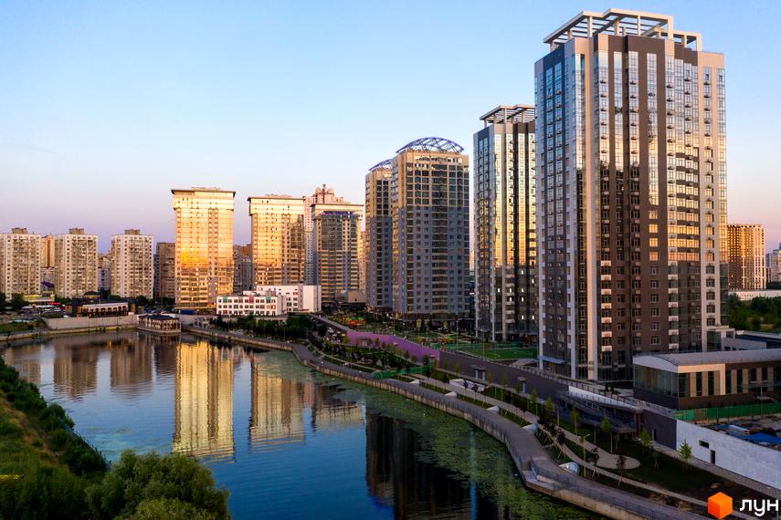 Житловий комплекс RiverStone – високі багатоповерхові будинки з дзеркальними фасадами, що відбивають сонячне світло. Набережна з пішохідними доріжками та зеленню.