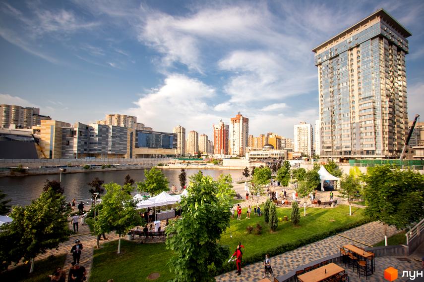 Житловий комплекс RiverStone з високою вежею та іншими багатоповерховими будинками, розташований на березі річки. На прибережній зоні облаштовано зелену зону з деревами, місцями для відпочинку та павільйонами.