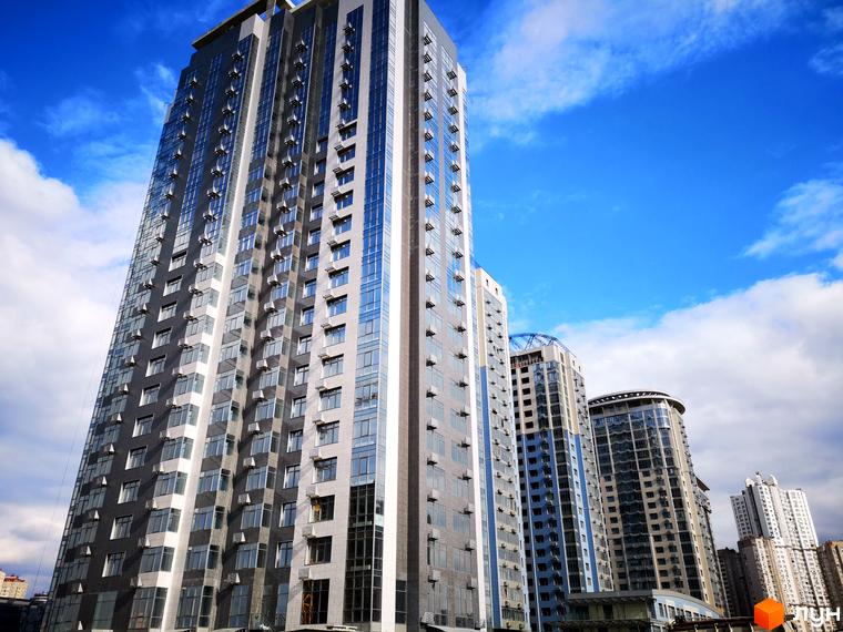 Житловий комплекс RiverStone - це сучасна архітектура висотних будинків з панорамними вікнами та скляними фасадами. Комплекс розташований на фоні блакитного неба з хмарами.