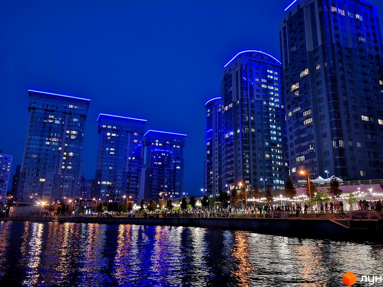Житловий комплекс RiverStone вночі. Багатоповерхові будинки з синім підсвічуванням фасаду, відображенням у воді. Набережна з людьми та деревами.