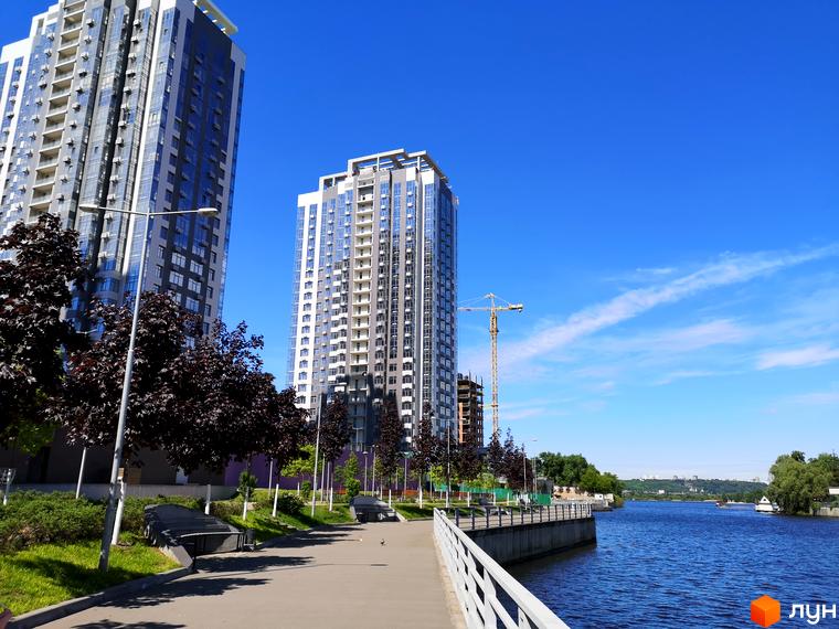 Житловий комплекс RiverStone з високими будинками та набережною. На території комплексу є зелені насадження та пішохідні доріжки.