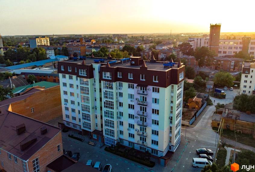 Зовнішній вигляд багатоповерхового будинку з балконами та кондиціонерами, прибудинкова територія з парковкою та зеленими насадженнями.