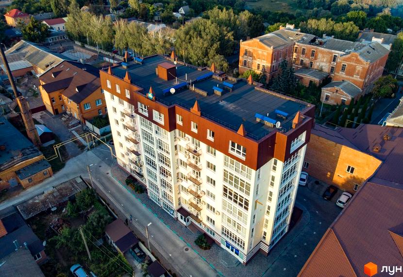 Зовнішній вигляд новобудови на вулиці Монастирська, 4г, з білим фасадом та коричневим дахом. На фото також видно прибудинкову територію з дорогою та деревами.