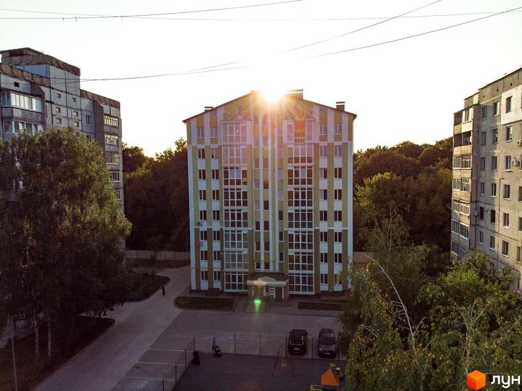 Будинок на вулиці Гостиннодвірська, 14в, з яскравим фасадом та великою кількістю вікон, на тлі дерев. Поруч розташована прибудинкова територія з автомобілями та дитячим майданчиком.