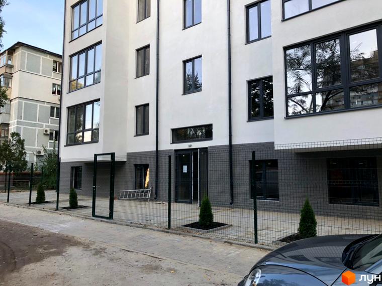 Зовнішній вигляд клубного будинку «Зразковий» з сітчастим парканом та зеленими насадженнями на прибудинковій території.